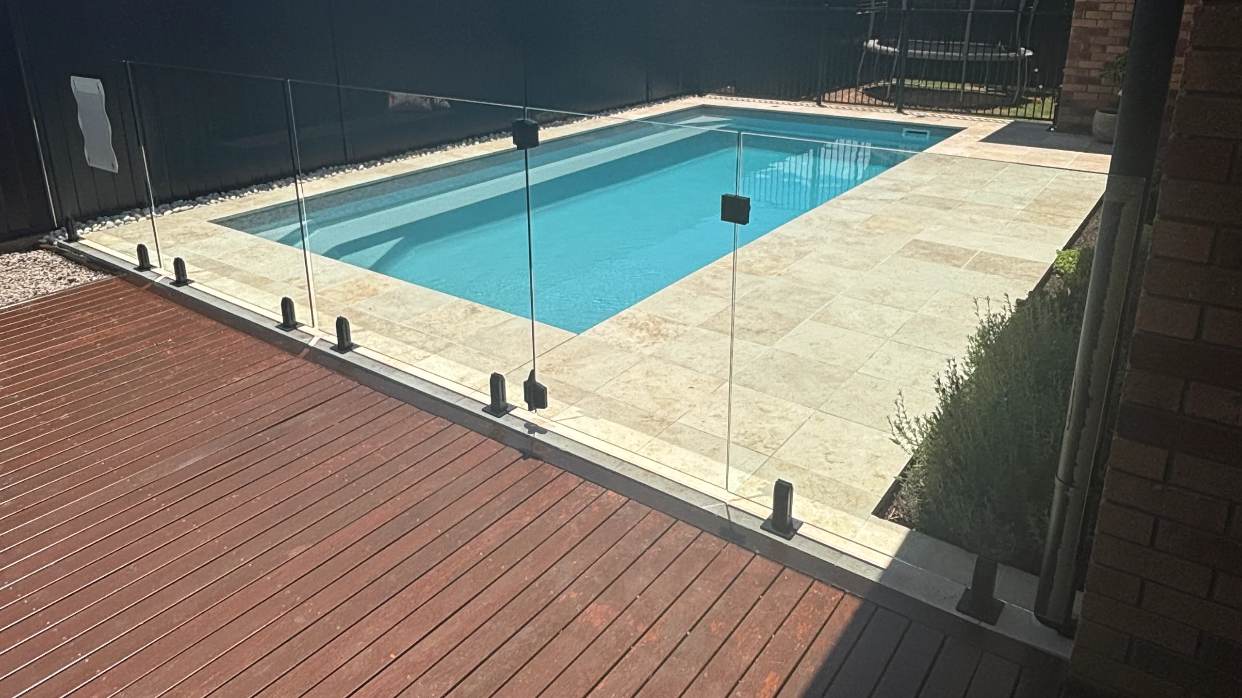 Rectangular fibreglass pool bordered by cream travertine pavers, timber decking and frameless glass pool fencing