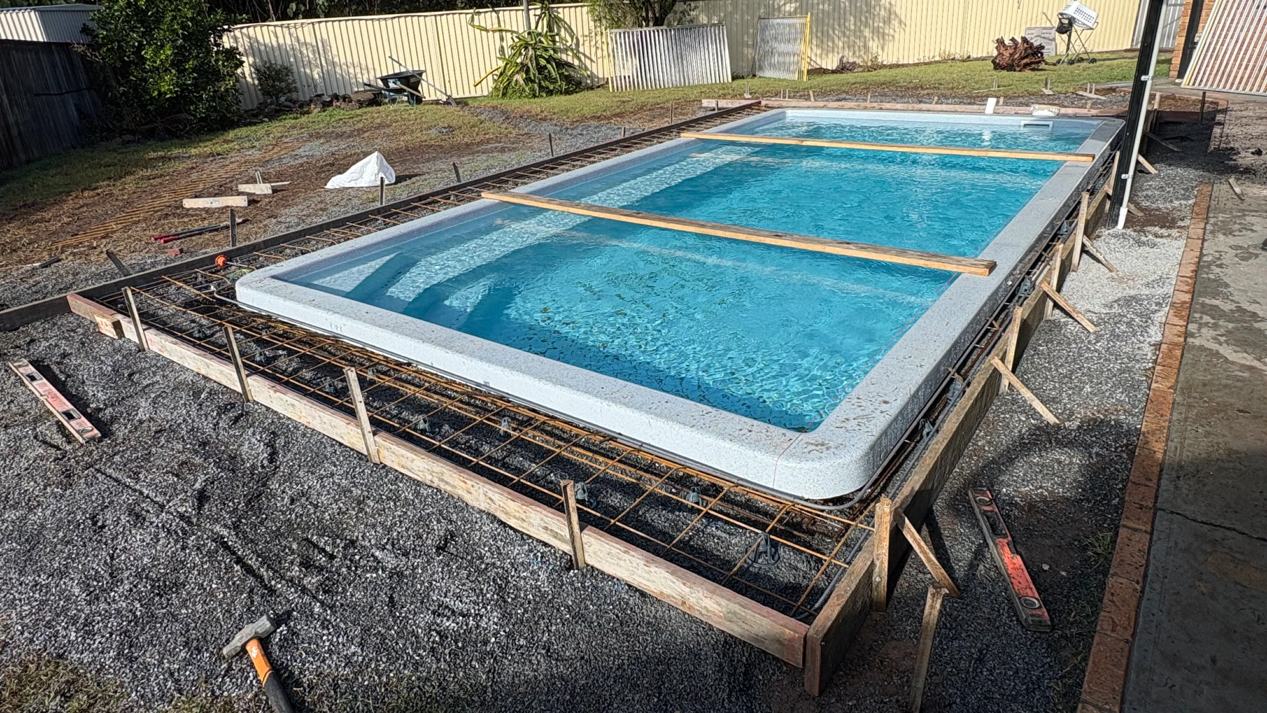Fibreglass inground pool filled with water sits in reinforced steel formwork awaiting concrete pour around the edges