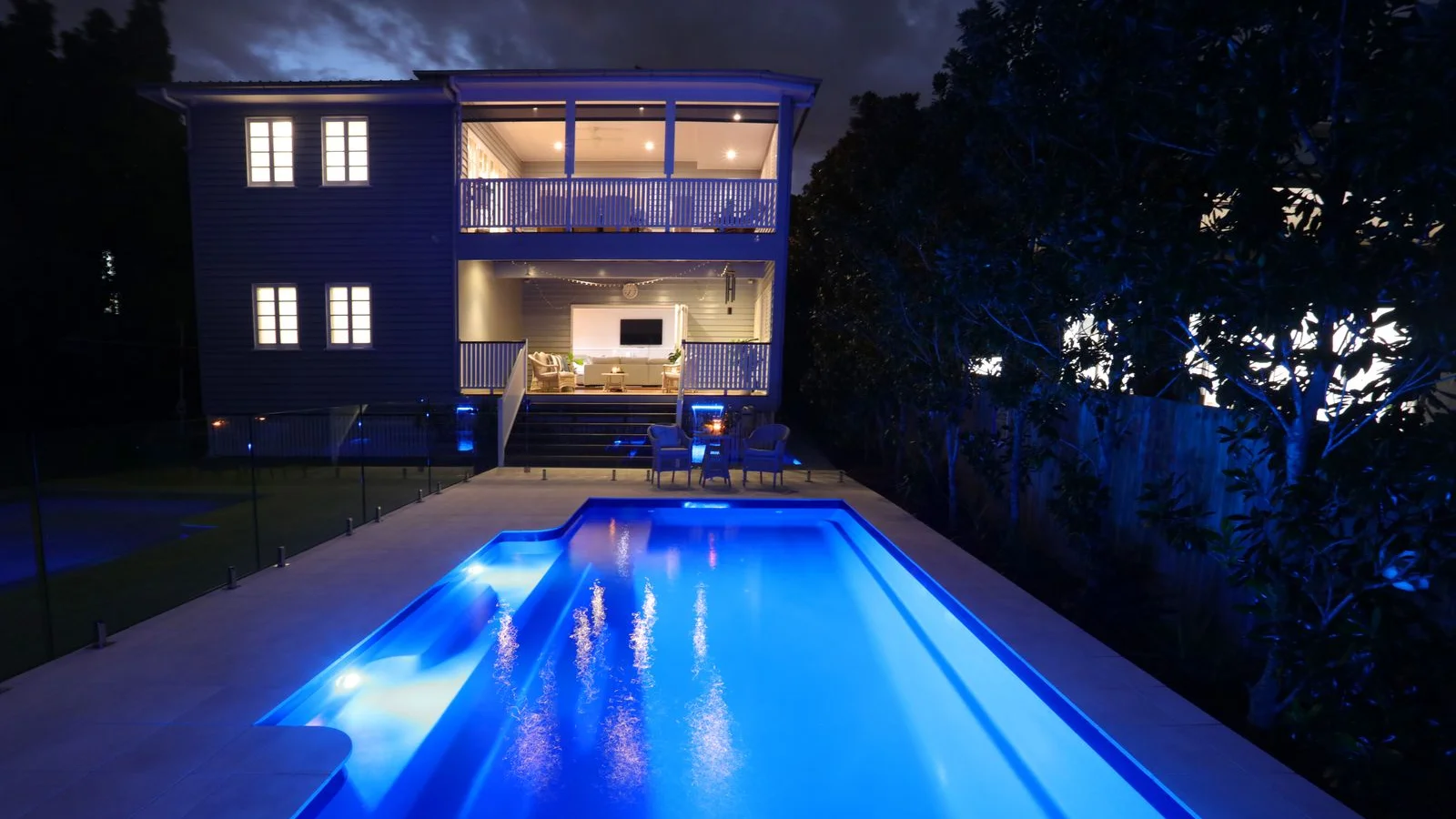 Blue LED lighting illuminates a fibreglass pool at night beside a two-storey home with a lit outdoor deck