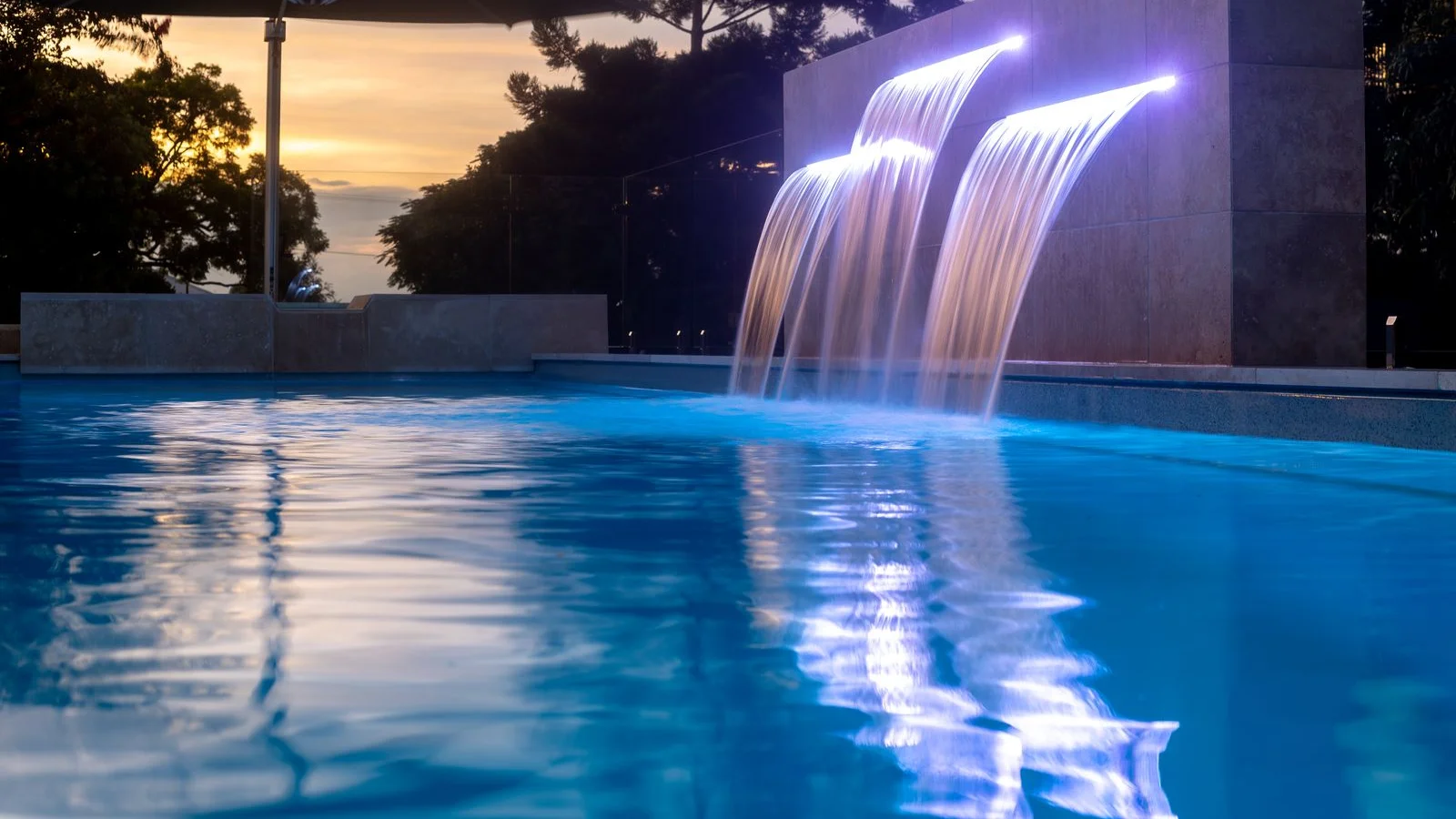 Three illuminated blade water features cascade into a blue fibreglass pool at dusk