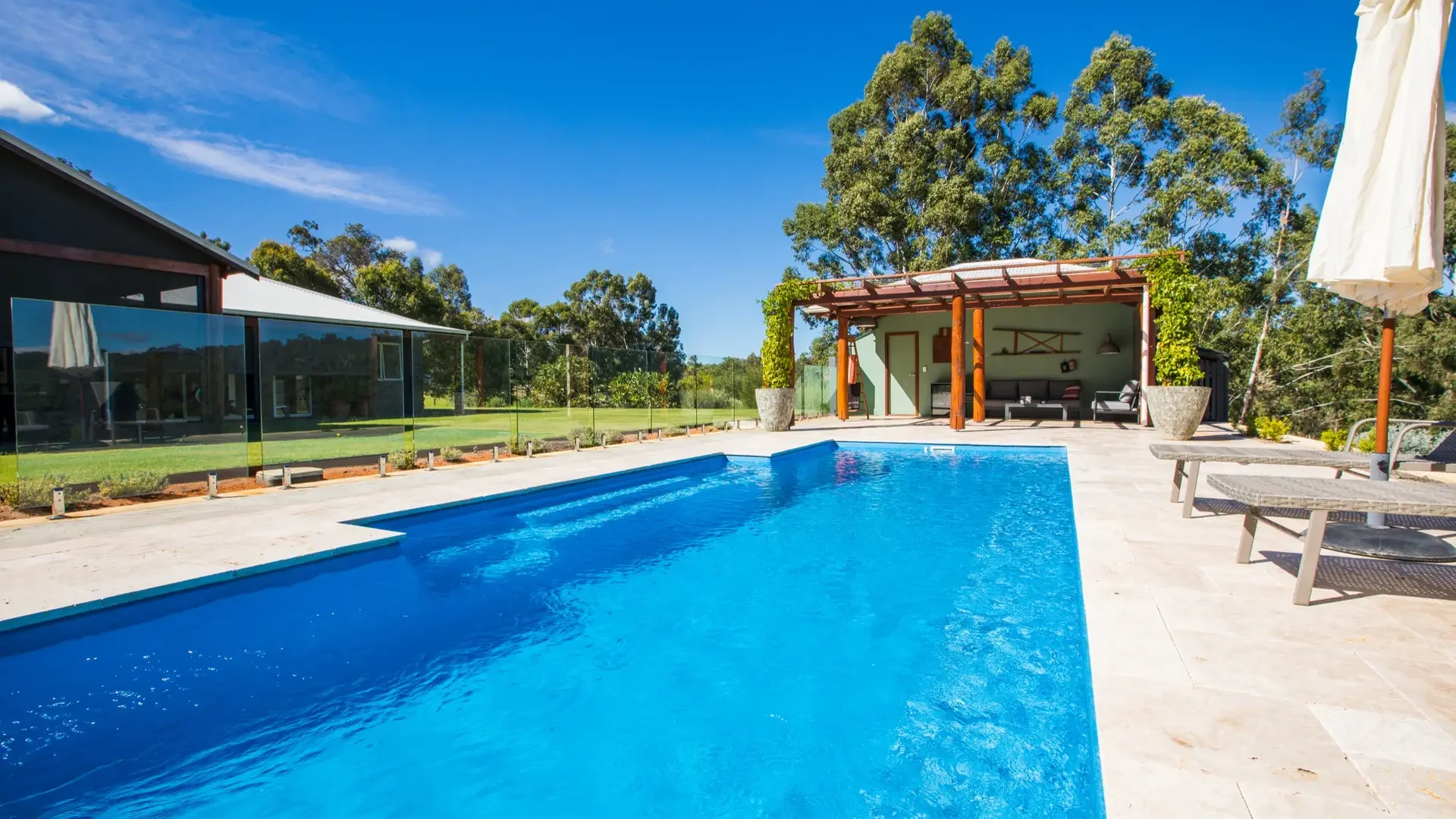 Rectangular fibreglass lap pool with cream travertine paving, glass fencing and a covered outdoor pavilion