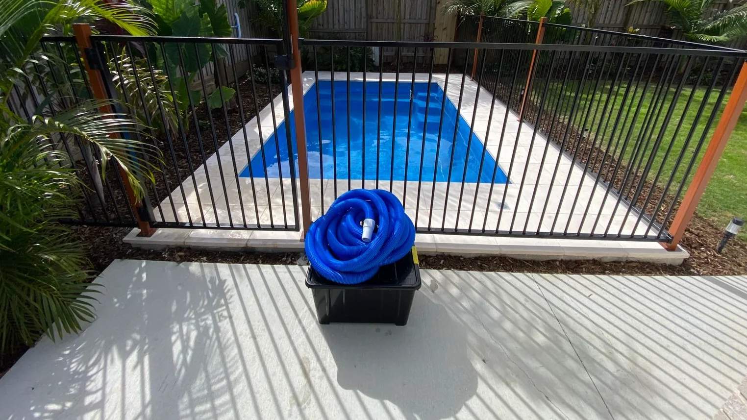 Coiled blue pool vacuum hose in a black storage tub beside a fibreglass pool behind black safety fencing
