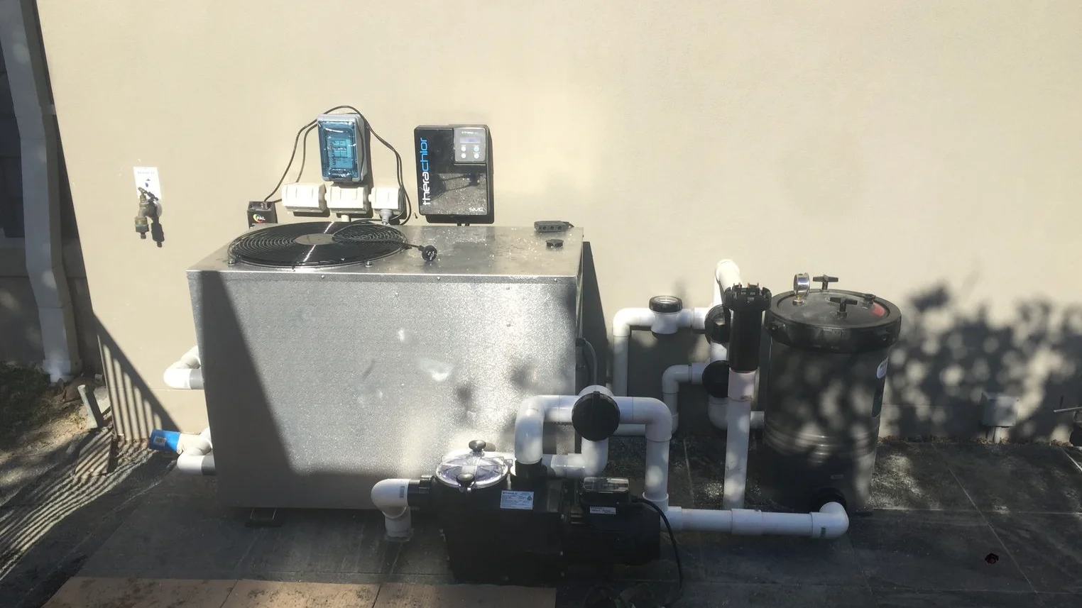 Pool heat pump with theraChlor chlorinator, pump and filter tanks connected by white PVC pipework against a wall