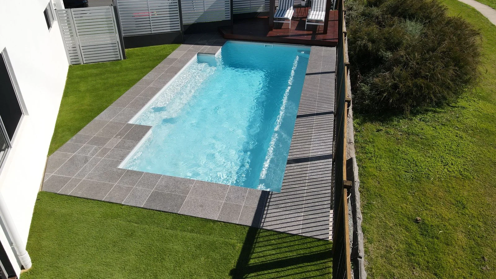 Rectangular fibreglass pool bordered by artificial turf, grey stone-look paving and composite decking