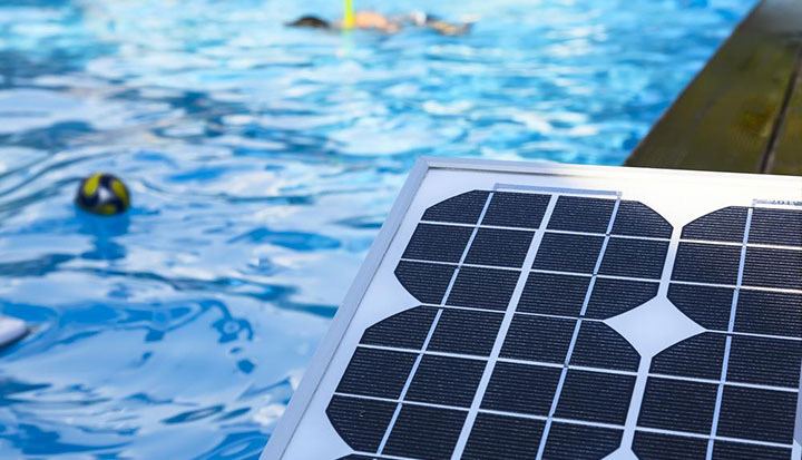 Solar pool heating panel with black collector cells resting beside a sunlit blue swimming pool