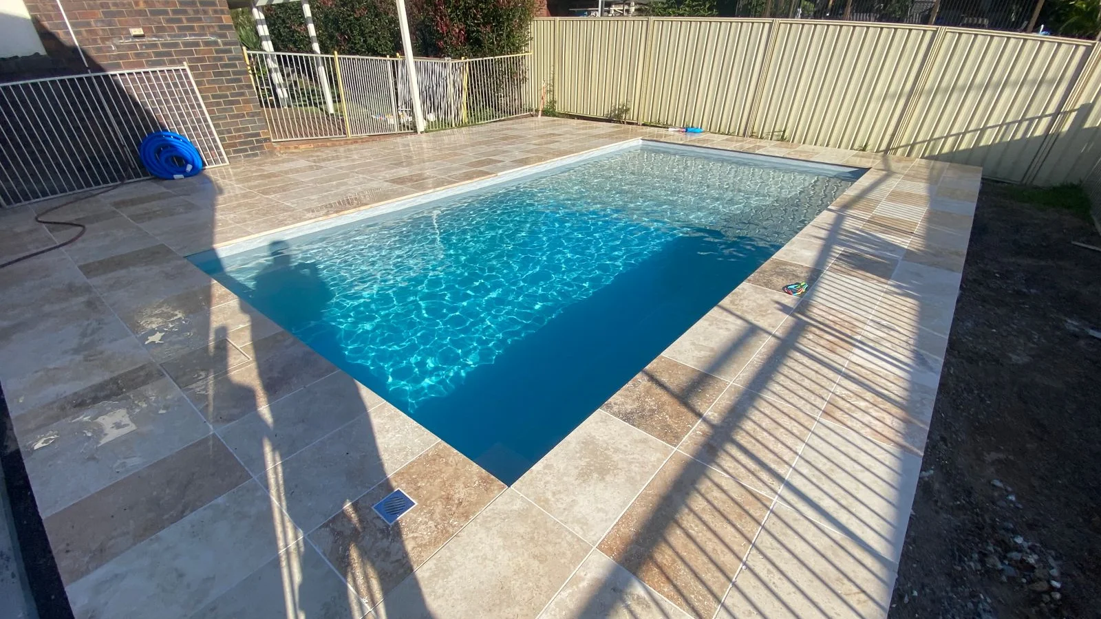 Rectangular fibreglass pool with a vacuum hose coiled on cream stone paving during pool cleaning