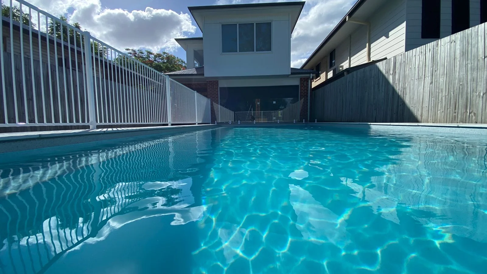 Fibreglass infinity pool with clear blue water bordered by white safety fencing and timber fence behind a two-storey home