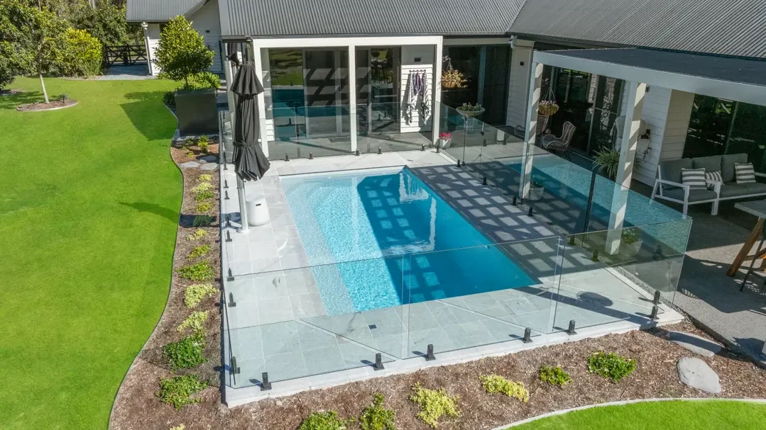 Glass pool fencing around a rectangular fibreglass pool with pale stone paving, lawn and garden beds beside a home