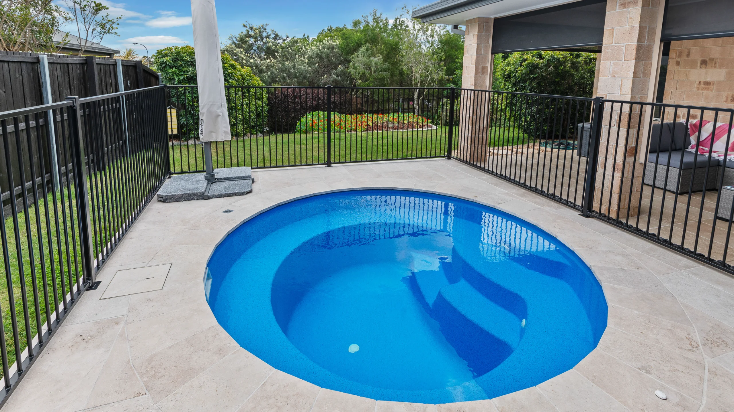 Round blue fibreglass magnesium pool set in cream stone paving with black fencing and garden