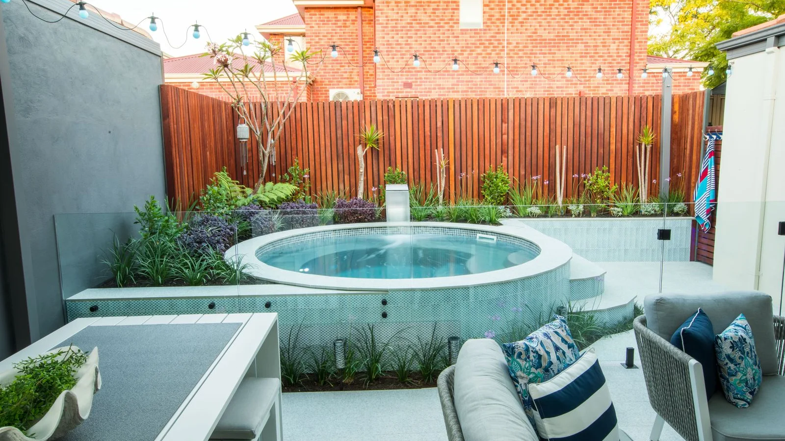 Round fibreglass plunge pool with water feature, glass fencing, timber screen and lush garden beds beside outdoor lounge seating