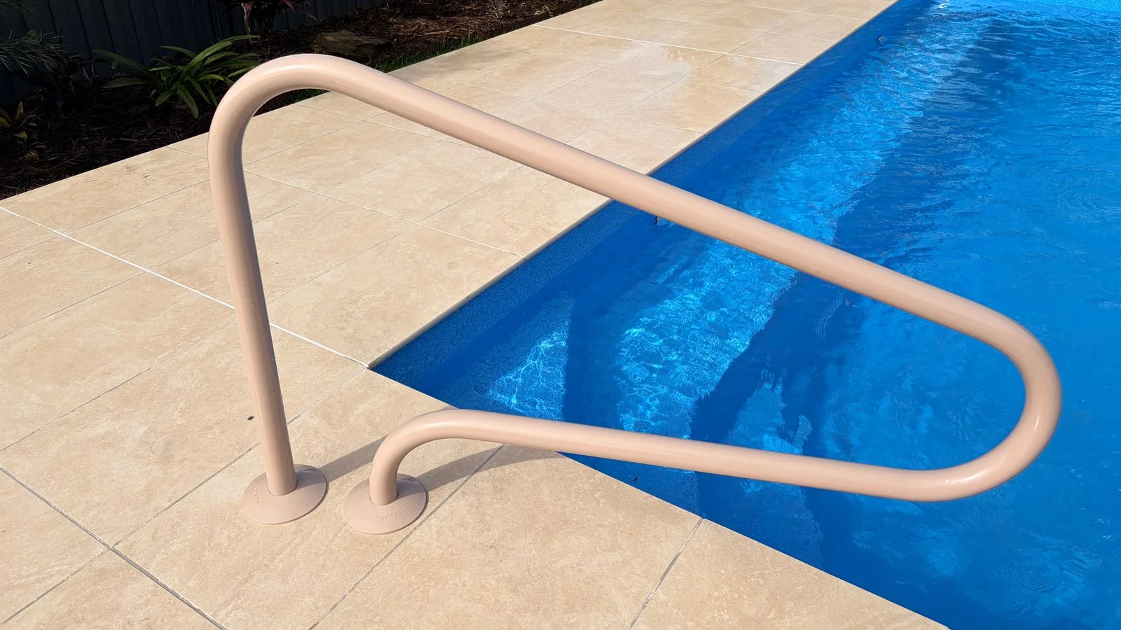 Beige pool entry handrail mounted on cream stone pavers beside a blue fibreglass pool