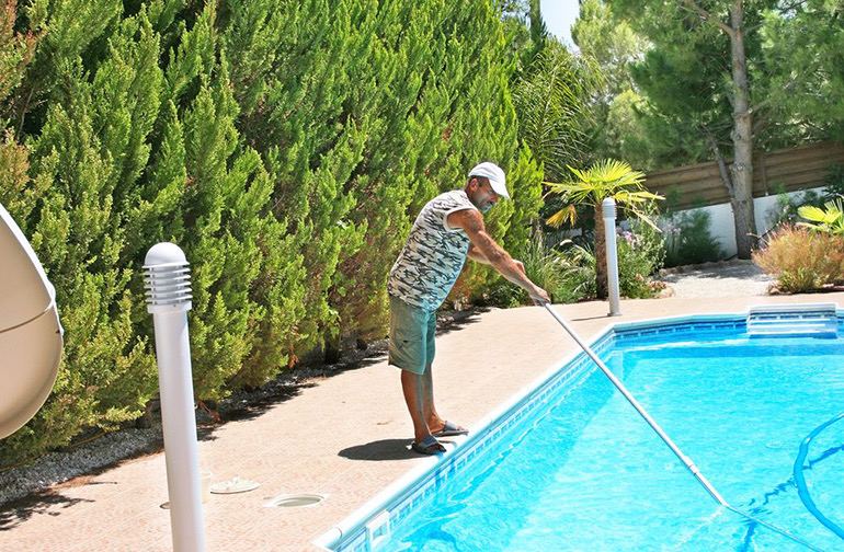 A man using a long-handled net to skim an inground fibreglass pool bordered by cream pavers and hedging