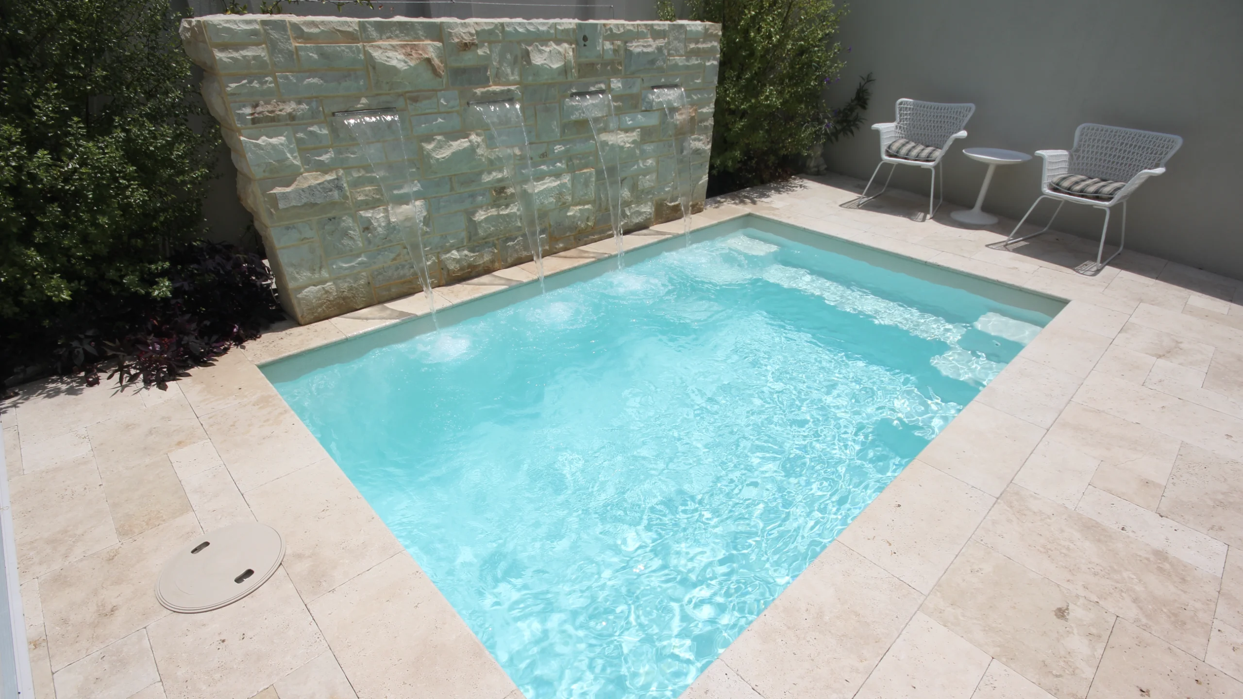 Compact rectangular fibreglass plunge pool with stone water wall and travertine paving in a small courtyard