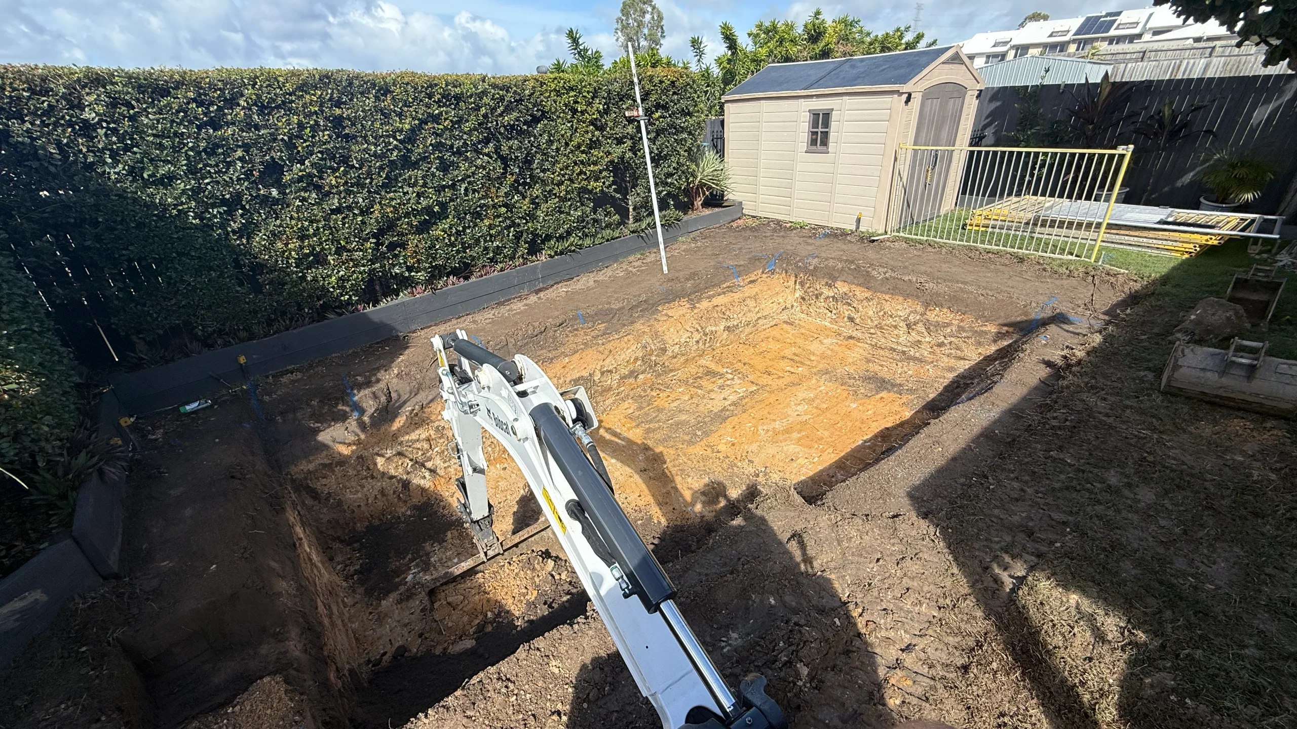 Mini excavator digging a rectangular inground pool hole in a backyard beside a hedge and garden shed