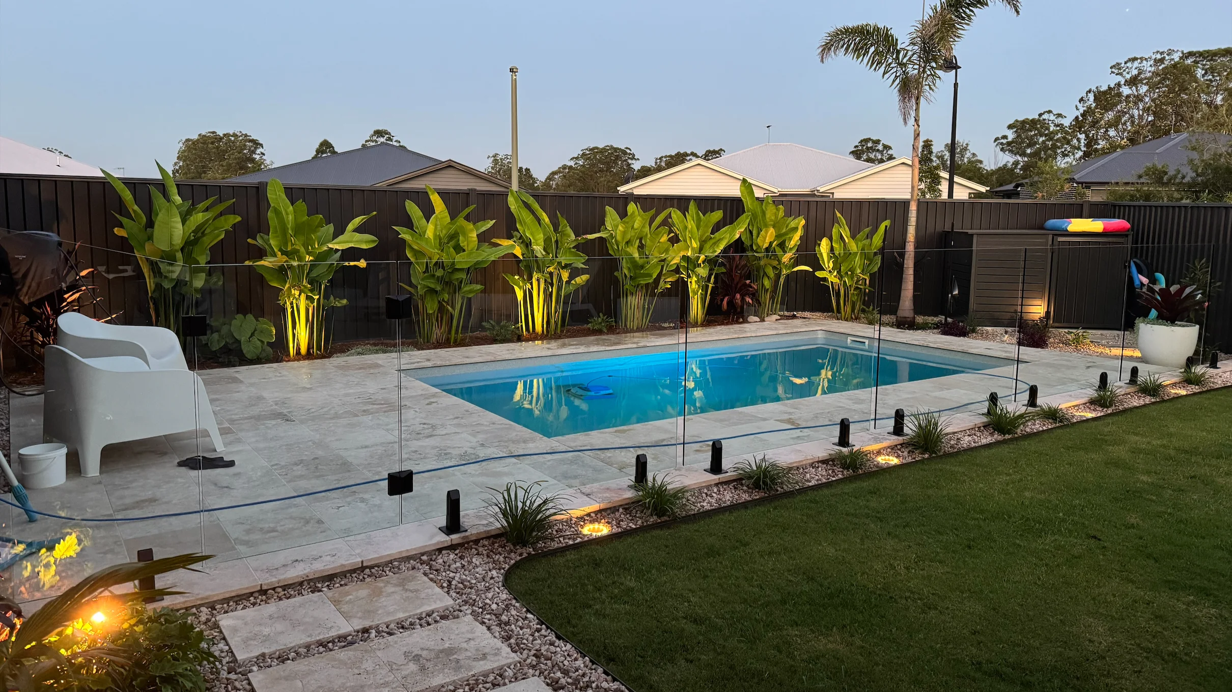Rectangular fibreglass pool framed by frameless glass fencing, travertine paving and lit tropical planting at dusk