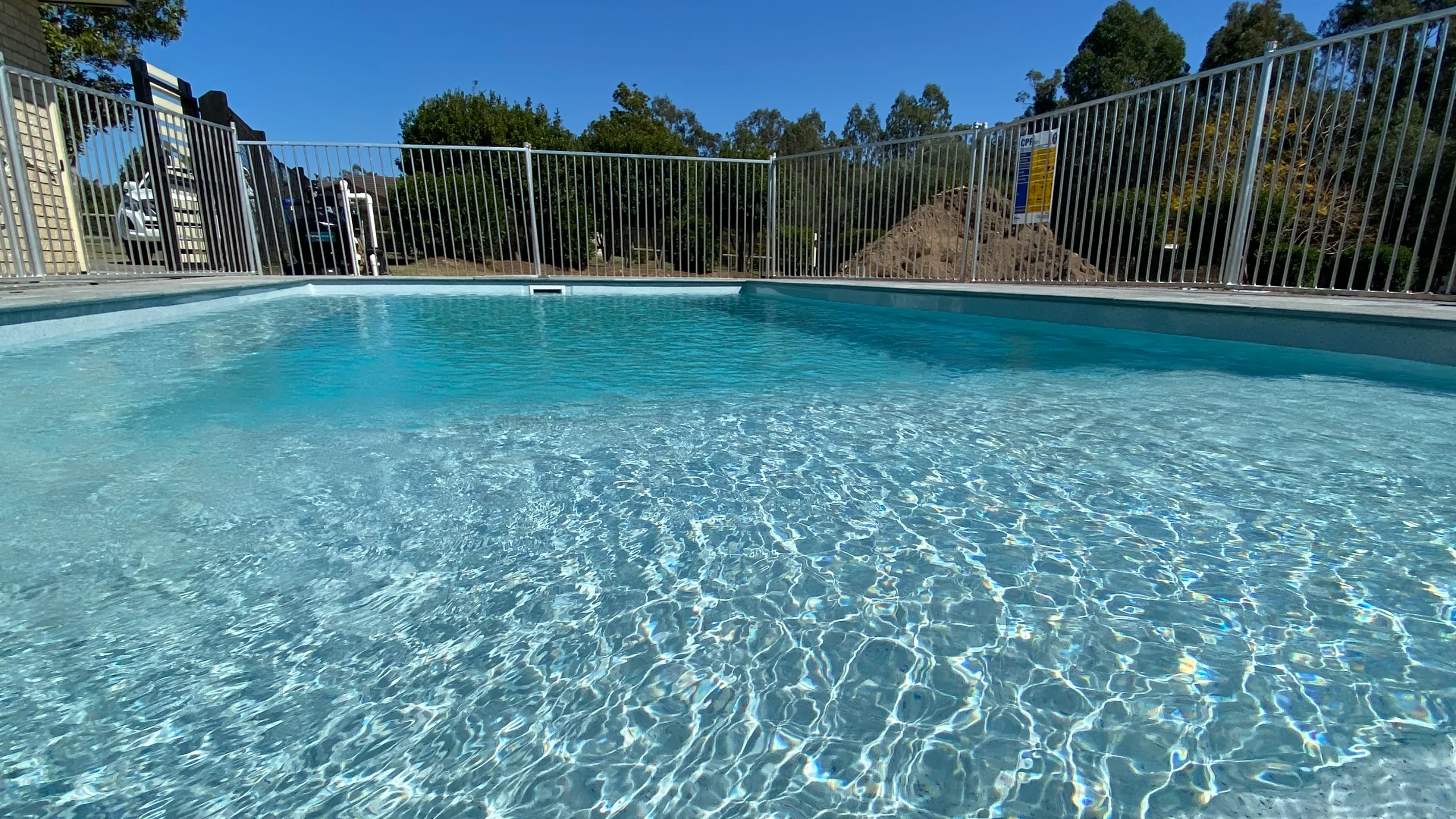 Crystal-clear fibreglass pool water rippling in sunlight behind a grey safety fence and trees