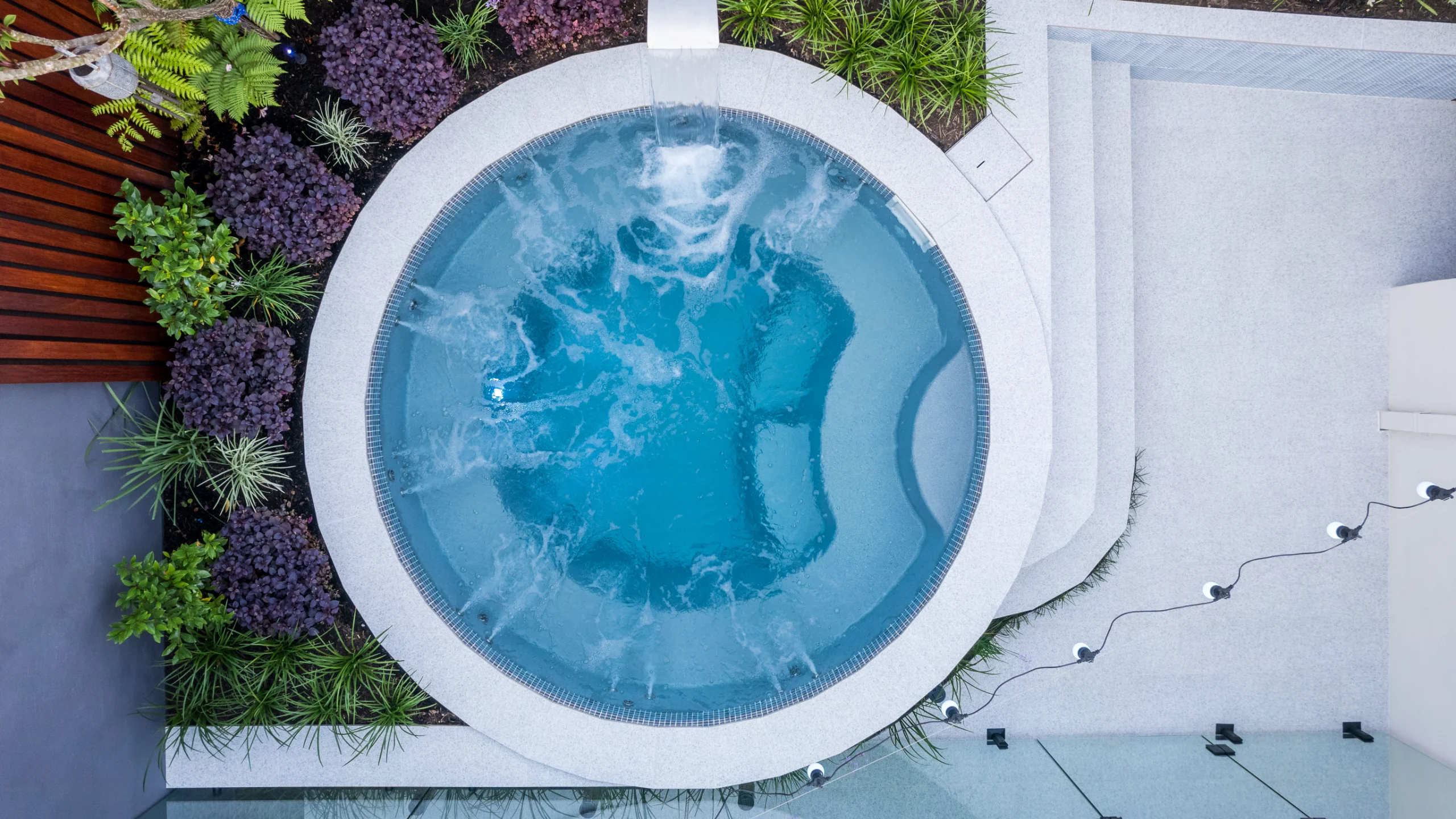 Round fibreglass spool pool with bubbling spa jets and a waterfall spout set in stone coping and lush garden