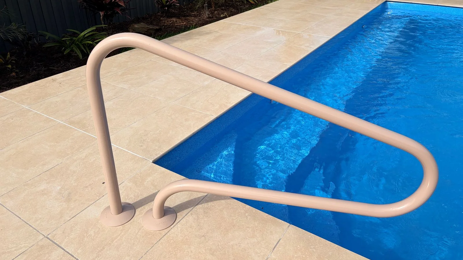 Beige powder-coated pool safety handrail beside cream paving and entry steps into a blue fibreglass pool