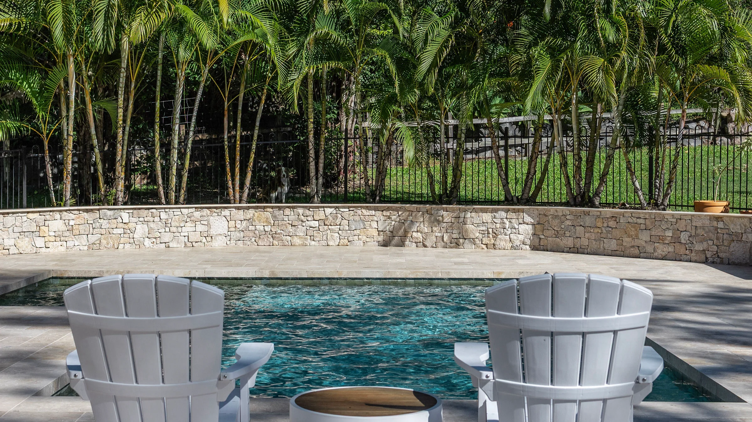 Fibreglass pool with clear blue water framed by white timber chairs, travertine paving and lush palm garden