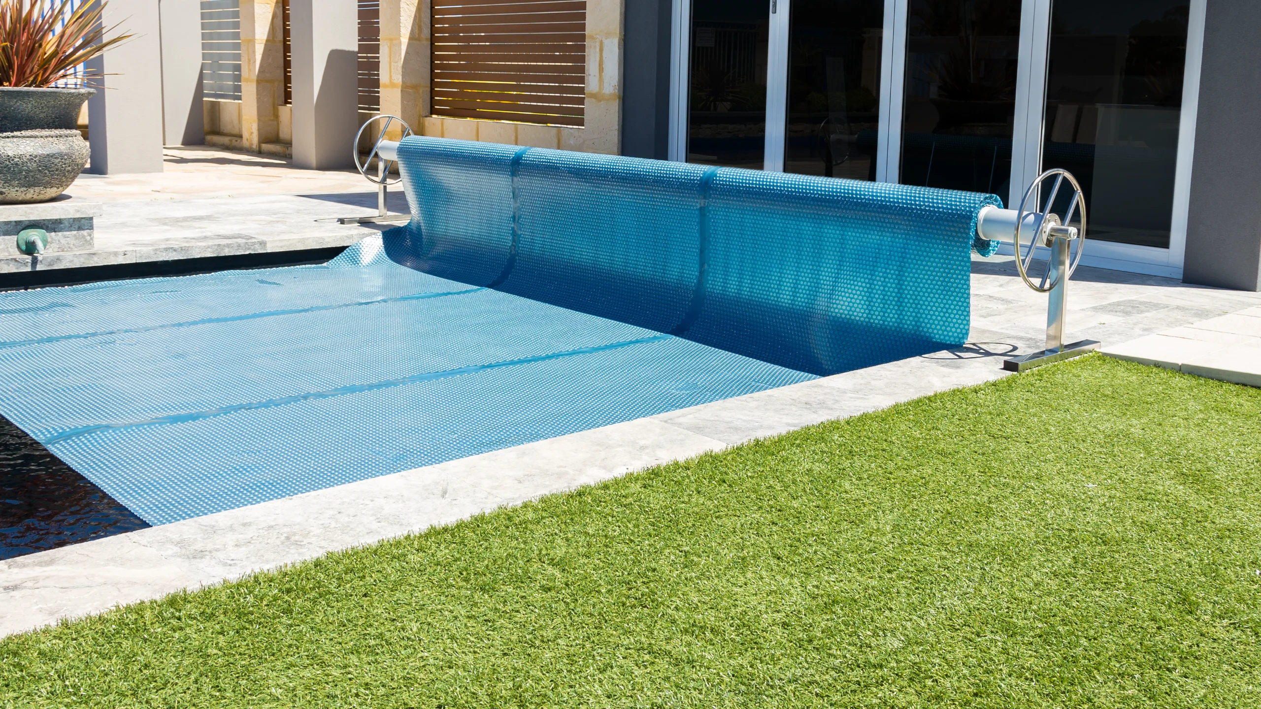 Blue bubble solar pool cover partly rolled onto a stainless steel reel beside a stone-edged pool