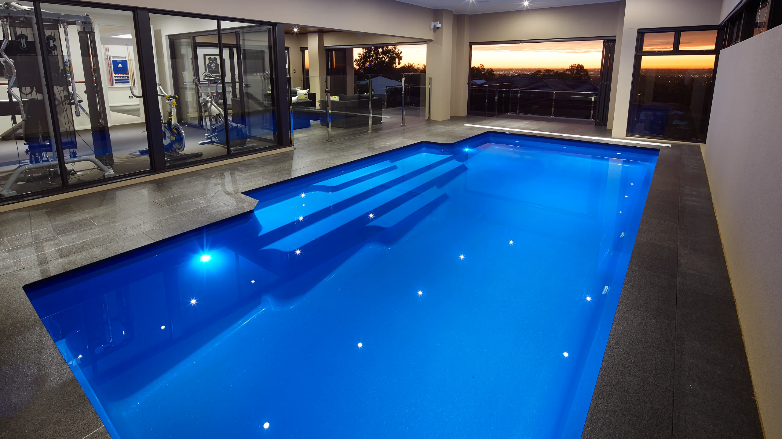 Indoor L-shaped fibreglass pool with blue LED lighting, dark stone paving and sunset city views beyond