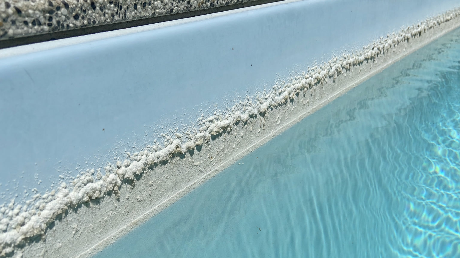 White crusty calcium scale band along the waterline tile line of a fibreglass pool above clear blue water
