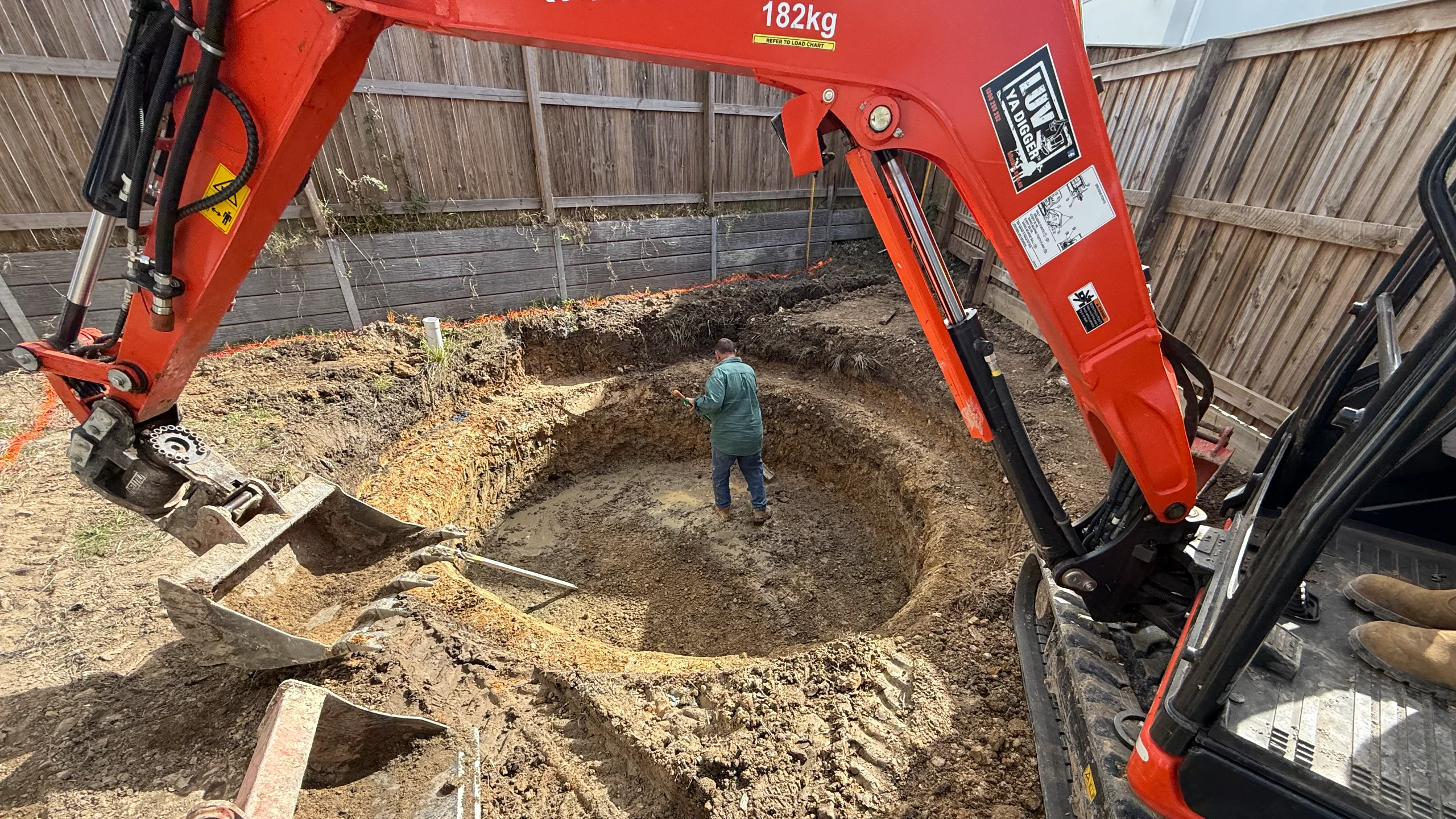 MFP Easy installer trimming a pool excavation while a red excavator digs the fibreglass shell hole in a fenced backyard