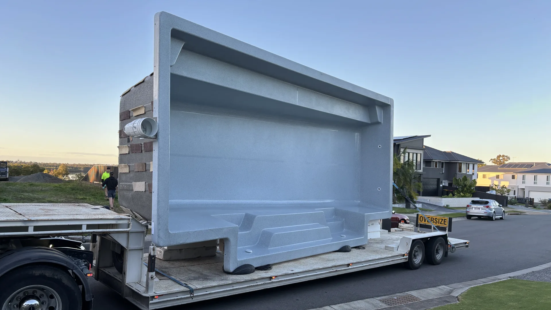 A one-piece fibreglass pool shell on its side on an oversize delivery trailer in a residential street