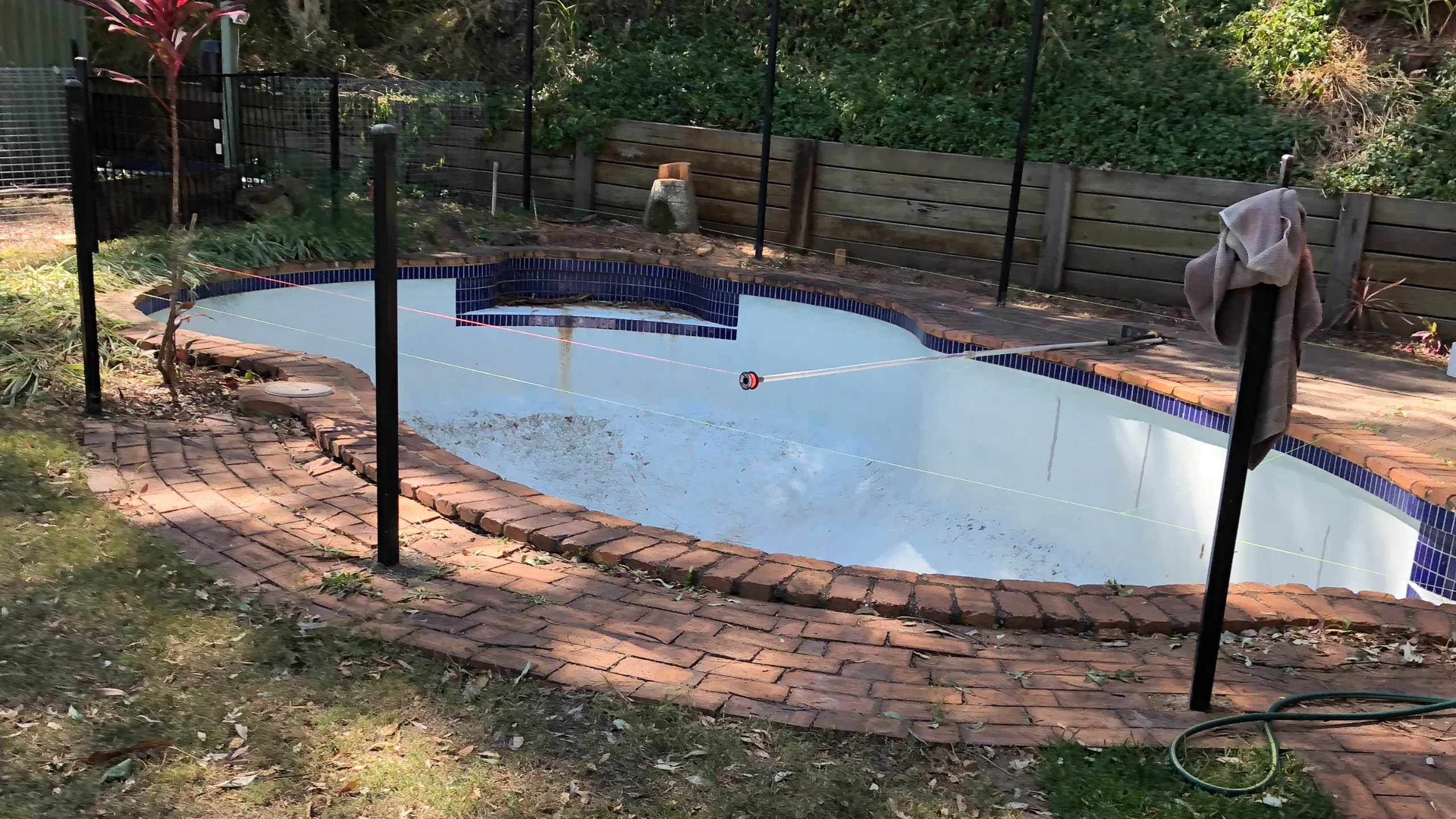Old drained pool ready for removal with tiled surrounds and brick coping in an overgrown backyard