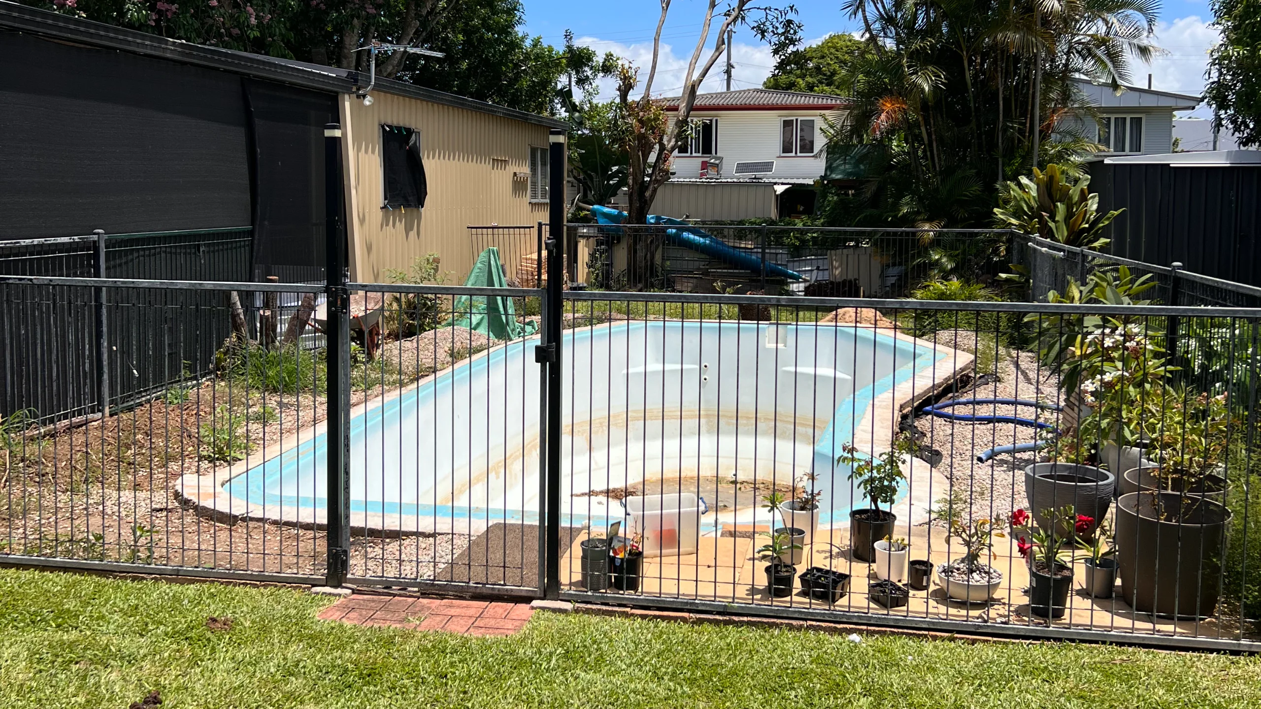 Empty fibreglass pool shell with faded, weathered gelcoat and staining behind a black safety fence