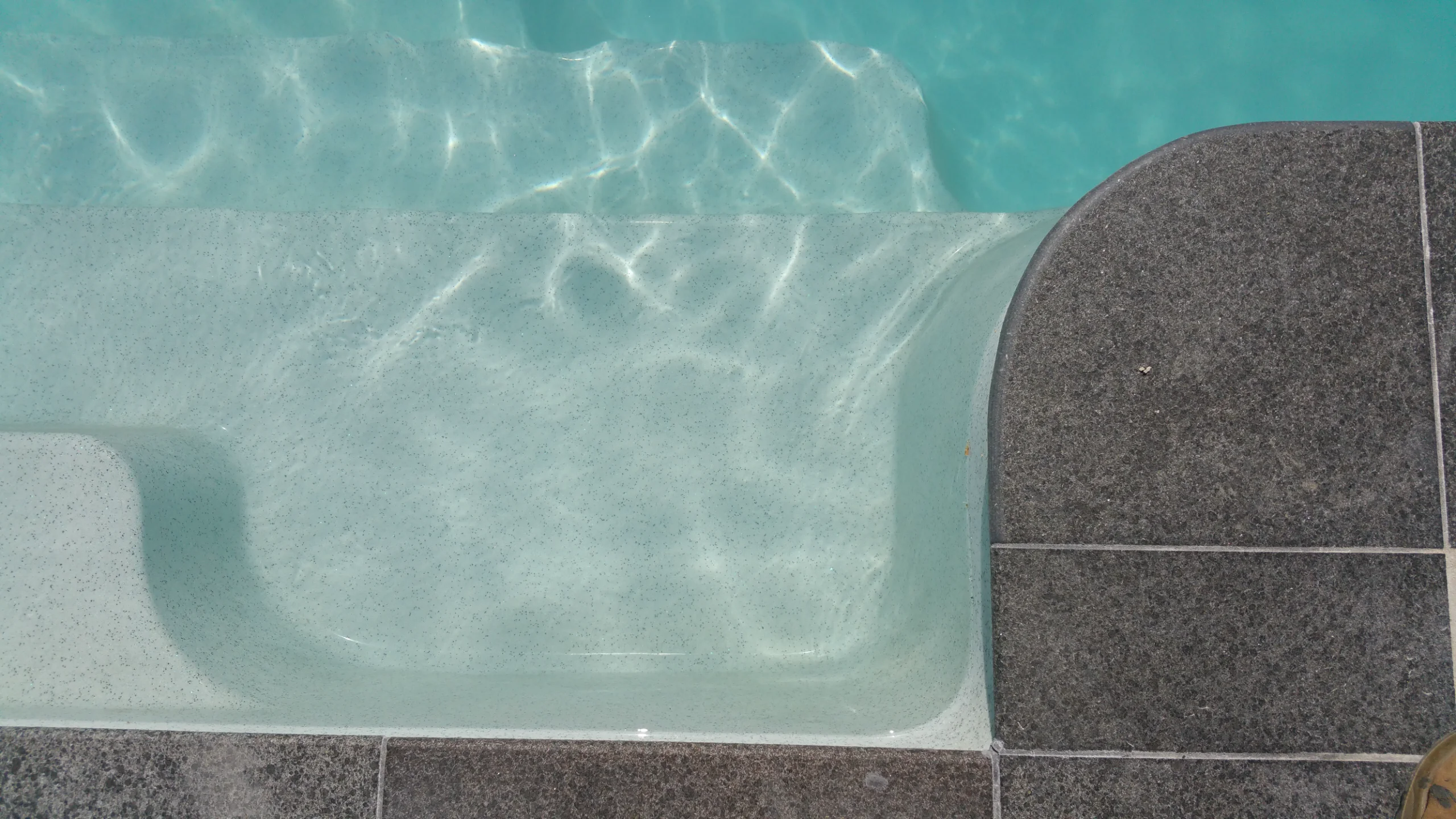 Pool paint on a fibreglass shell step and bench beside grey granite coping tiles at the water's edge